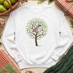 May include: A white long-sleeve sweatshirt featuring a central graphic of a brown and grey partridge bird perched in a stylized tree with green leaves and small green pears. The design includes a small text label at the base of the tree trunk.