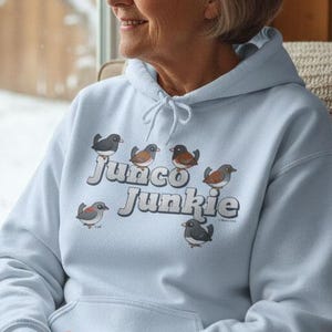 May include: A light blue hoodie with the text "Junco Junkie" and illustrations of small birds. The hoodie has a drawstring and is made of a soft material. The person is sitting in a chair near a window.