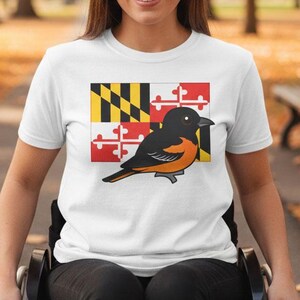 May include: White t-shirt featuring a graphic of a Maryland flag and an orange and black bird. The shirt is a short-sleeved crew neck style. The background shows a park with trees and a bench.