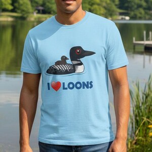 May include: A light blue t-shirt featuring a graphic of a loon with its chick. The text "I ❤️ LOONS" is printed below the illustration. The shirt is worn by a person standing in front of a lake.