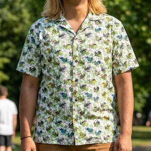 May include: A short-sleeved button-down shirt with a white background and a repeating pattern of green, blue, and yellow birds. The shirt is paired with brown pants. The person is smiling and standing outside in a park.