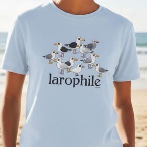 May include: Light blue t-shirt featuring a cartoon illustration of several seagulls. The word "larophile" is printed in a stylized font below the birds. The shirt is made of a soft material and is suitable for casual wear.