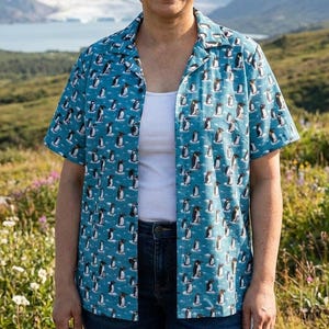 May include: A short-sleeved, button-down shirt in a light blue color, patterned with numerous penguin illustrations. The shirt has a classic collar and is worn open over a white tank top and blue jeans. The background features a scenic landscape.