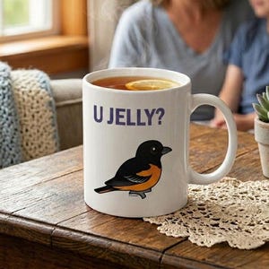 May include: White ceramic mug with the text "U JELLY?" and a cartoon bird illustration. The bird is black and orange. The mug is filled with tea and a lemon slice.