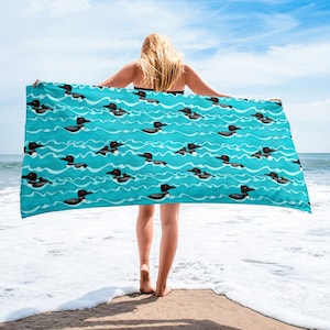 May include: A turquoise beach towel featuring a repeating pattern of black and white loons swimming on wavy lines. The towel is held up by a person on a sandy beach with the ocean in the background.