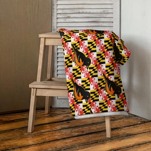 May include: A rectangular throw blanket featuring a repeating pattern of the Maryland state flag in red, white, black, and yellow, interspersed with illustrations of black and orange Baltimore orioles perched throughout the fabric design.