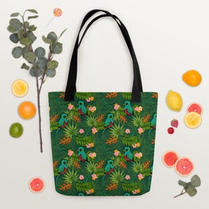 May include: A tote bag featuring a repeating pattern of green quetzal birds, tropical palm leaves, and small pink and orange flowers on a dark green background. The bag has two black fabric handles attached to the top edge of the rectangular body.