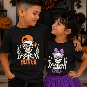 May include: Two black t-shirts featuring a skeleton graphic with the text "COUSIN CREW" on sunglasses. The left shirt shows a skeleton in an orange baseball cap with the name "OLIVER" below. The right shirt shows a skeleton in a purple bow with "EMILY".