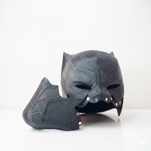 Puede incluir: Un casco negro de Pantera Negra con una placa frontal desmontable. El casco tiene una superficie texturizada con detalles intrincados y remaches plateados. La placa frontal presenta un diseño geométrico y una sección perforada.