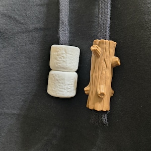 May include: Two dark gray hoodie drawstrings featuring decorative aglets. One drawstring has two white, textured marshmallow-shaped beads stacked vertically, while the other features a brown, cylindrical log-shaped bead with detailed bark texture.