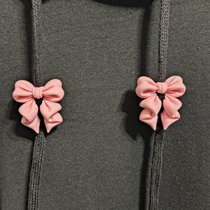 Puede incluir: Una sudadera con capucha de color gris oscuro que presenta dos lazos de color rosa claro a juego sujetos a los dos cordones verticales. Los lazos están colocados simétricamente en la parte delantera de la prenda, cada uno unido a un cordón.