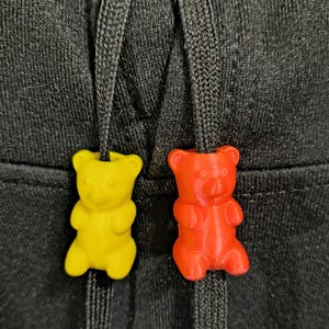 以下が含まれることがあります： 黒いドローストリングのクローズアップ。2つのグミベア型のコードエンドが付いています。1つは黄色、もう1つは赤です。クマは光沢のある仕上げで、黒い衣服のドローストリングに取り付けられています。