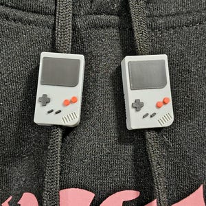 May include: Two white and gray Game Boy-style charms with black and orange details. The charms are attached to a black hooded sweatshirt. The charms have a retro video game aesthetic.