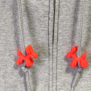 May include: A heather grey zip-up hoodie featuring two bright orange balloon dog-shaped aglet covers attached to the grey braided drawstrings. The hoodie has a central metal zip and a textured fabric surface with visible knit details.