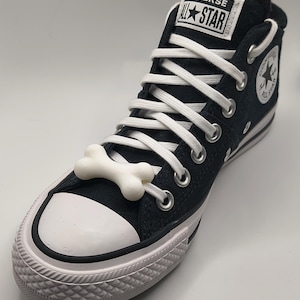 May include: Black Converse All Star high-top sneaker with white laces and a white rubber toe cap. A white bone-shaped charm is laced into the shoe. The Converse All Star logo is visible on the tongue and side of the shoe.