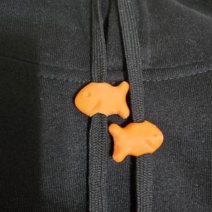 Könnte beinhalten: Nahaufnahme eines schwarzen Kapuzenpullovers mit zwei orangefarbenen Goldfisch-Crackern, die als Kordelstopper verwendet werden. Die Cracker sind leuchtend orange und haben ein lächelndes Gesicht.