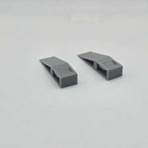 以下が含まれることがあります： 2つのグレーの3Dプリントドアストッパー。各ストッパーは、傾斜した上部と長方形の切り欠きを備えた長方形のベースを備えています。ドアストッパーは、ドアが閉まるのを防ぐように設計されています。