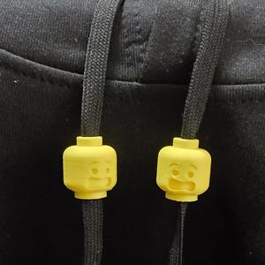 Könnte beinhalten: Zwei gelbe, Lego-ähnliche Kopf-Kordelenden an einem schwarzen Kapuzenpullover-Kordelzug. Jeder Kopf hat einen anderen Gesichtsausdruck, einer lächelt und der andere ist überrascht. Der Kapuzenpullover ist schwarz.