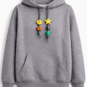 Puede incluir: Sudadera con capucha gris con bolsillo de canguro y capucha con cordón. Los cordones tienen adornos decorativos de colores: un cero amarillo, un cuadrado marrón, una estrella amarilla y cilindros verdes y negros. Una sudadera informal y cómoda.