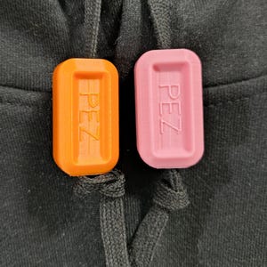 May include: Close-up of two rectangular PEZ-branded charms, one orange and one pink, resting on a black fabric. The charms are 3D printed and have the word "PEZ" embossed on them. The charms are attached to a black drawstring.