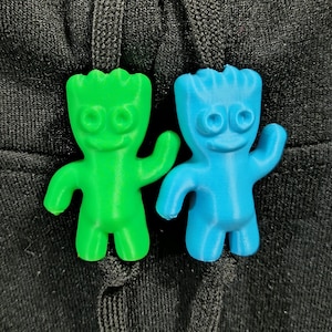 May include: Two small, colorful, 3D-printed figures in the shape of a smiling character. One is bright green, the other is light blue. They have large eyes, arms raised, and a wavy head. The figures are on a black, textured surface.