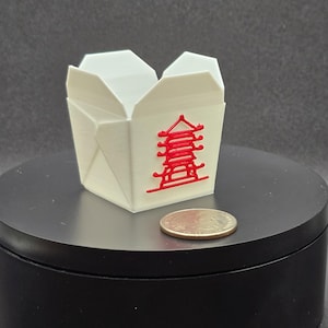 May include: A small, white takeout container with a red pagoda design. The container is open at the top and sits on a black surface. A U.S. quarter is next to the container for size comparison.