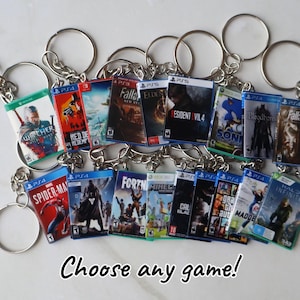 May include: A collection of miniature video game case keychains featuring various titles such as The Witcher, Resident Evil 4, Spider-Man, and Halo Infinite. Each keychain has a metal ring and chain attached to a small rectangular game cover replica.