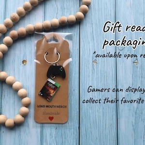 May include: A keychain featuring a black video game controller charm and a miniature Fallout game case replica attached to a silver metal ring, mounted on a brown cardboard display card labeled LOUD MOUTH MERCH Handmade with a red heart.
