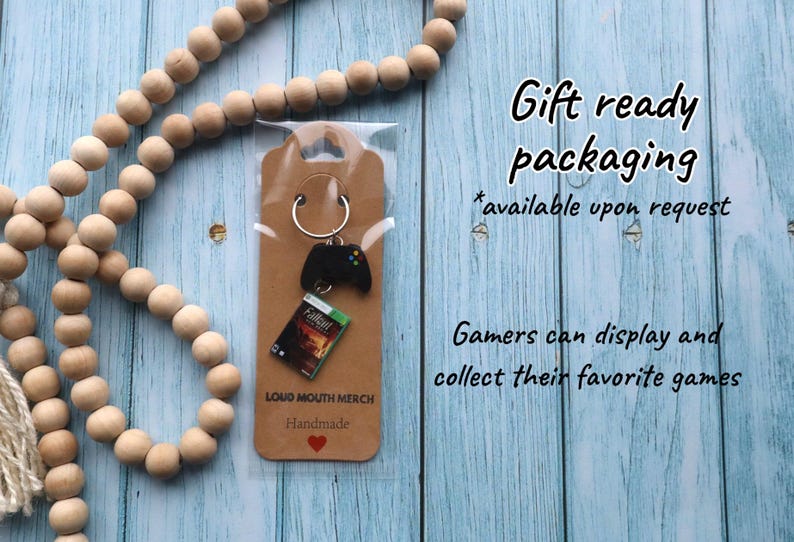 May include: A keychain featuring a black video game controller charm and a miniature Fallout game case replica attached to a silver metal ring, mounted on a brown cardboard display card labeled LOUD MOUTH MERCH Handmade with a red heart.