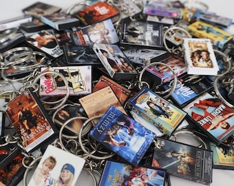 May include: A collection of miniature rectangular keychains featuring various movie poster designs on the front. Each keychain is attached to a silver metal split ring. The movie titles visible include My Cousin Vinny, The Last of Us, and Nacho Libre.