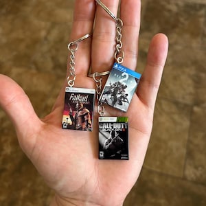 May include: Three miniature video game case keychains attached to a silver metal ring. The keychains feature the cover art for Fallout New Vegas for Windows, Destiny 2 for PS4, and Call of Duty Black Ops II for Xbox 360, each with a small metal chain.