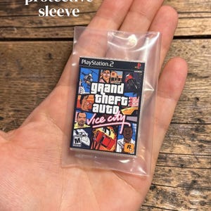 May include: A miniature replica of the Grand Theft Auto Vice City video game case for the PlayStation 2 console, featuring the iconic multi-panel cover art with characters, vehicles, and the game title in pink script, enclosed in a clear plastic sleeve.