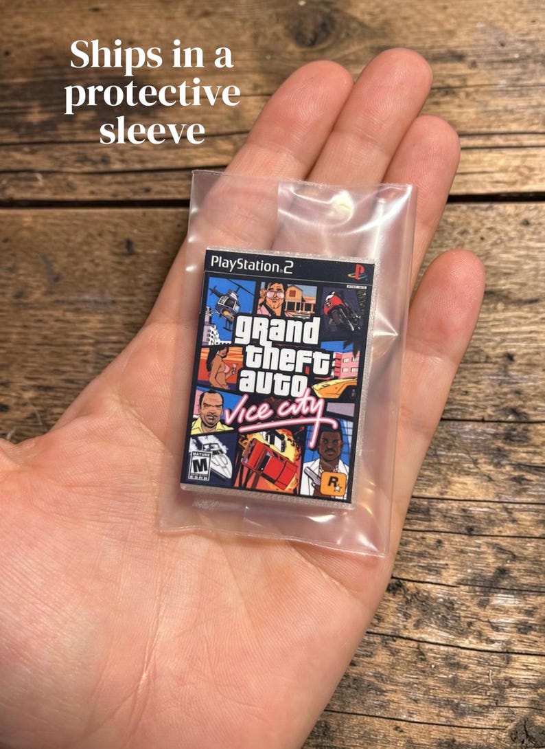 May include: A miniature replica of the Grand Theft Auto Vice City video game case for the PlayStation 2 console, featuring the iconic multi-panel cover art with characters, vehicles, and the game title in pink script, enclosed in a clear plastic sleeve.