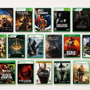May include: A collection of fifteen video game cases arranged in three rows. Titles include Age of Empires, Gears of War 2, Arc Raiders, Fight Night Champion, Black Myth Wukong, Gears of War 3, Elden Ring, Assassin's Creed, Crimson Desert, Red Dead Redemption 2, Halo, Red Dead Redemption, Gears of War, The Witcher 3 Wild Hunt, Call of Duty Modern Warfare Remastered, and Call of Duty Modern Warfare 2.