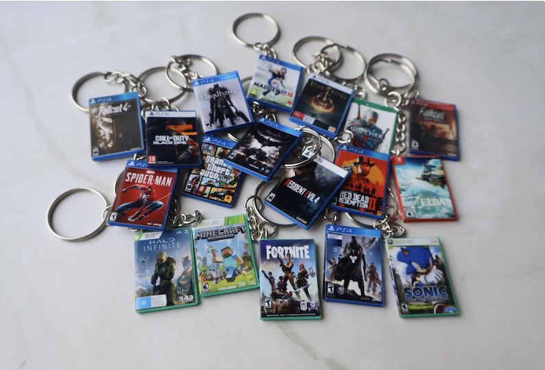 May include: A collection of miniature rectangular keychains featuring various video game cover art, including titles like Spider-Man, Minecraft, Fortnite, and Halo Infinite. Each keychain has a metal ring attached to a small plastic case.