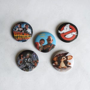 May include: Five circular pin-back buttons featuring 1980s movie imagery. Designs include the Back to the Future logo, the Ghostbusters symbol, characters from The Goonies with the text Hey you guys!, Beetlejuice, and the creature Gizmo from Gremlins.