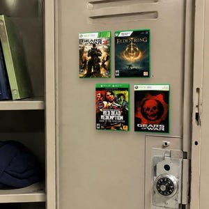 May include: Four video game cases attached to a beige metal locker door. The titles include Gears of War 3, Elden Ring, Red Dead Redemption Game of the Year Edition, and Gears of War. Each case features its respective cover art and console branding.