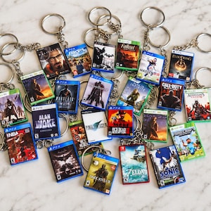 May include: A collection of miniature rectangular keychains featuring various video game cover art designs. Each keychain includes a metal split ring and chain, displaying titles such as Elden Ring, Minecraft, Zelda, and Red Dead Redemption 2.