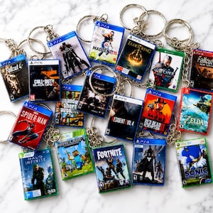 May include: A collection of miniature video game case keychains featuring various titles including Fallout 4, Call of Duty Black Ops III, Bloodborne, Madden 15, Elden Ring, The Witcher, Fallout New Vegas, Spider-Man, GTA, Resident Evil 4, Red Dead Redemption II, Zelda, Halo Infinite, Minecraft, Fortnite, Destiny, and Sonic.