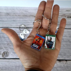 May include: Two miniature rectangular keychains featuring video game cover art. One keychain displays the red and orange Red Dead Redemption 2 artwork with a PS4 logo, while the other shows The Witcher 3 Wild Hunt with a green Xbox One logo.