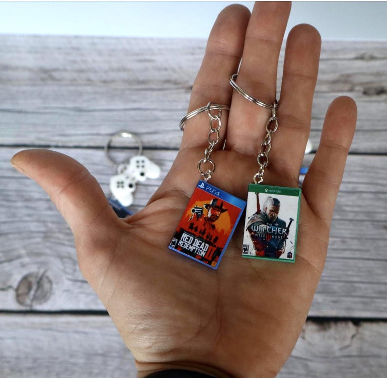 May include: Two miniature rectangular keychains featuring video game cover art. One keychain displays the red and orange Red Dead Redemption 2 artwork with a PS4 logo, while the other shows The Witcher 3 Wild Hunt with a green Xbox One logo.
