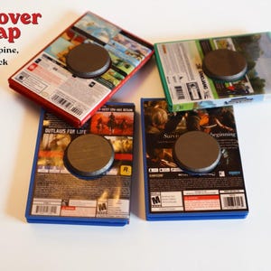 May include: Four rectangular video game cases with full cover wraps featuring front, spine, and back artwork. Each case has a circular dark gray magnet attached to the center of the back cover. The cases are arranged in a cluster on a plain surface.