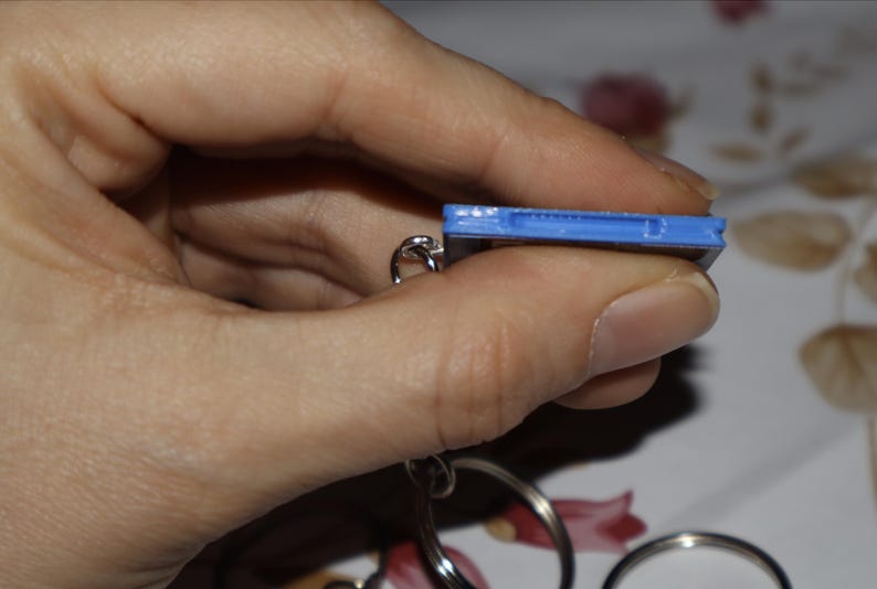 May include: A blue rectangular plastic keychain attachment featuring a thin slot along the top edge, connected to a circular silver metal split ring. The object is shown from a side profile view, highlighting the blue casing and the metal hardware.