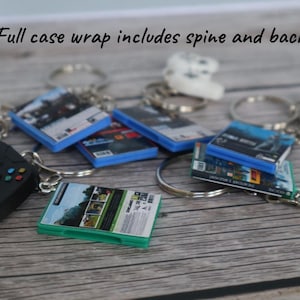 May include: A collection of miniature video game case keychains featuring full case wraps with spines and backs, accompanied by small black and white game controller charms attached to silver metal key rings. The text reads: Full case wrap includes spine and back.
