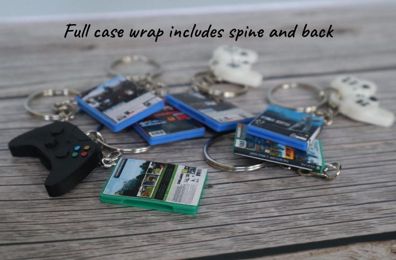 May include: A collection of miniature video game case keychains featuring full case wraps with spines and backs, accompanied by small black and white game controller charms attached to silver metal key rings. The text reads: Full case wrap includes spine and back.
