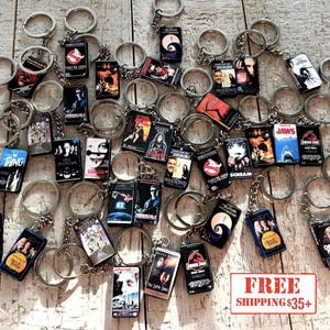 May include: A collection of movie-themed keychains featuring various film titles. Each keychain has a small, rectangular design with movie posters and attached to a silver keyring. The keychains are scattered on a wooden surface.