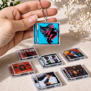May include: A collection of miniature square plastic CD case keychains featuring various album cover art, including Pearl Jam, TLC CrazySexyCool, Gorillaz, and Michael Jackson Thriller. Each clear case has a metal ball chain attachment.