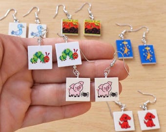Op de afbeelding: Een collectie zilverkleurige oorhangers met vierkante witte bedels voorzien van kleurrijke illustraties van dieren en insecten, waaronder een groene rups, een roze varken met een spin, een blauwe vogel, een gele giraf en een rode hond.