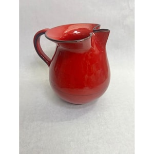 May include: A glossy, vibrant red ceramic pitcher featuring a rounded, bulbous body, a curved handle on the left side, and a distinctively shaped pouring spout with a wavy, undulating rim edge that is finished with a thin, dark trim.