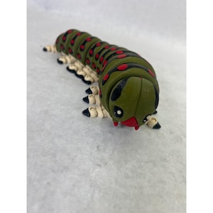 May include: An articulated caterpillar figurine featuring an olive green body with black stripes and red spots. The creature has a rounded head with large white eyes, a red mouth area, and multiple beige legs with black tips along its segmented body.